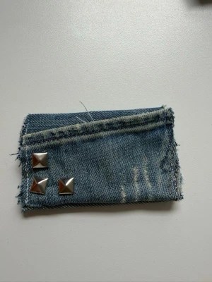 Jeansplånbok med nitar - Cool plånbok i blått denimtyg med slitna detaljer och tre silverfärgade pyramidnitar på framsidan. Plånboken har en rå och edgy look med fransiga kanter och är perfekt för dig som gillar streetstyle och unika accessoarer.