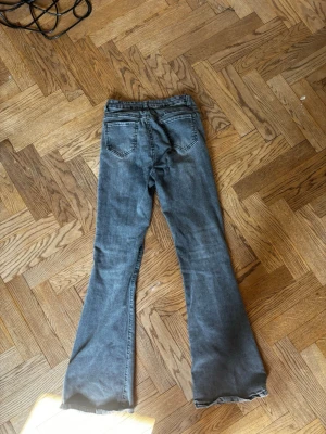 Grå bootcut jeans med rå kant - Säljer ett par grå bootcut jeans med rå, fransad kant nertill. Jeansen har klassiska bakfickor och en snygg tvättad look. Perfekta för dig som gillar en avslappnad och trendig stil.