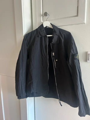 Svart overshirt från Stone Island - Svart overshirt från Stone Island med klassisk krage och dragkedja framtill. Jackan har lång ärm och ikonisk logotyp på vänster ärm. Tillverkad i nylon. Nypris är 5400kr.