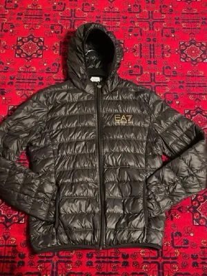 Svart pufferjacka från EA7 Emporio Armani - Säljer en svart pufferjacka från EA7 Emporio Armani med huva och dragkedja framtill. Jackan har quiltad design, elastiska muddar och EA7-logga i guld på bröstet. Perfekt för kalla dagar och riktigt snygg streetstil.