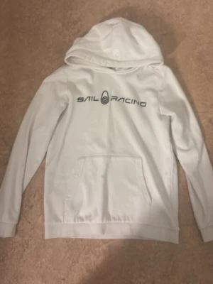 Vit hoodie från Sail Racing - Vit hoodie från Sail Racing med svart logga tryckt över bröstet. Klassisk känguruficka framtill och huva. Perfekt för dig som gillar en clean och sportig stil. Mjuk bomullskänsla och långa ärmar. den har en liten fläck på fickan och en liten fläck på luvan men knappt något som syns 