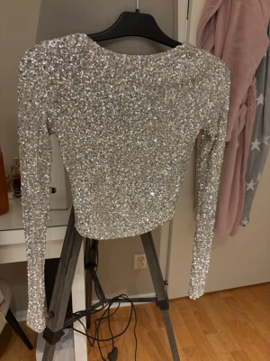 Glittrig silver topp med öppen rygg - Superglittrig långärmad topp i silver med paljetter över hela tyget. Toppen är croppad och har en snygg öppen rygg med en liten knäppning bak. Perfekt för dig som vill sticka ut och glänsa lite extra.