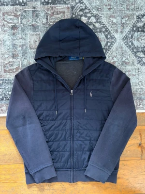 Mörkblå Ralph Lauren Cardigan/Hybrid zip Hoodie - Hej! 👋 Säljer en riktigt snygg quiltad Ralph Lauren hybrid zip jacka / cardigan i storlek M. Sjukt stilrent lyxplagg! Tröjan är i gott skick överlag. Den är lite sliten på ena ärmen samt vid ärmsluten, och kan upplevas som något solblekt på ryggen (inget som stör, se bilder). Priset är satt därefter! 100% äkta med originaltvättlappar kvar. Qr-kod finns! ✅Perfekt övergångsjacka eller lyxigare basplagg! Fraktar snabbt! 💬💯🚛📦 