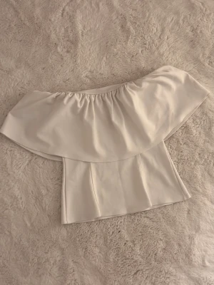 Vit offshoulder topp - Superfin vit offshoulder topp med bred volang runt axlarna. Snygg och stilren modell som sitter tajt över kroppen och har en croppad passform. Toppen är aldrig använd och har bara legat i garderoben, den passar också en större S eller mindre L🌸🩷