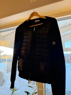 Svart cardiganjacka från Moncler - Svart cardiganjacka från Moncler med quiltad front och stickade ärmar. Jackan har dragkedja framtill och Moncler-logga på ärmen. Snygg och stilren design som passar perfekt till lager-på-lager-look.
