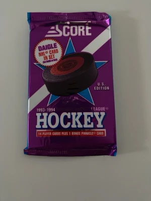 Score NHL Hockey kort 1993-94 - Ett oöppnat paket Score NHL Hockey samlarkort från säsongen 1993-1994, U.S. Edition. Förpackningen är lila med blå och vita detaljer och innehåller 14 spelarkort samt 1 bonus Pinnacle-kort. Perfekt för dig som gillar hockey och samlarkort.