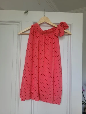 Rosa prickig topp med rosett - Supersöt coral topp med svarta prickar och en stor rosett på ena axeln. Toppen är ärmlös, har en loose passform och är gjord i ett lätt, luftigt syntetmaterial. Perfekt för dig som gillar färg och vill sticka ut med en lekfull detalj. I halterneck modell och såååp trendig och fin! Älskar färgen men den kommer tyvärr inte till användning och behöver platsen I garderoben! Tunntarmen material med den är linad så ej genomskinlig!