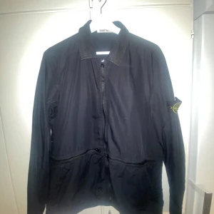 Äkta Svart vindjacka från Stone Island - Svart overshirt från Stone Island med klassisk krage och dragkedja framtill. Jackan har två fickor med dragkedja och den.  Perfekt för dig som gillar stilrena och funktionella plagg.