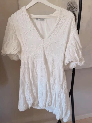 Vit kort klänning med puffärm från Zara - Superfin vit kort klänning från Zara med v-ringning och puffärmar. Klänningen har broderade detaljer och en lätt volang nertill. Perfekt för sommaren, även perfekt för studenten! Är i stl L men skulle säga mer M.  Använd endast en gång för att testa. 