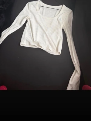 Vit ribbad långärmad crop top - Snygg vit långärmad crop top i ribbat material med rundad urringning. Toppen har en tight passform och är perfekt att styla med jeans eller kjol. Materialet är stretchigt och bekvämt, vilket gör den enkel att bära hela dagen.