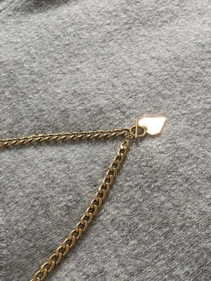 Guldfärgat halsband med hjärtberlock - Säljer ett chunky guldfärgat halsband med en platt hjärtformad berlock och en cool toggle-låsning. Kedjan är bred och har en modern look som verkligen sticker ut. Perfekt för dig som gillar statement-smycken och vill ha något unikt.