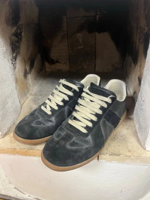 Svarta sneakers med mockadetaljer - Margielas som jag köpte här på plick. Lite trasig i hälen men syns och känns ej. (Vet ej om de är äkta det får ni avgöra).                                       Stilrena svarta sneakers med ovandel i både mocka och läder, vita snören och beige sula. Klassisk låg modell med retrovibbar och kontrasterande material. Perfekta för dig som gillar en clean och tidlös look. Jag är osäker på om det är äkta margellas eftersom 