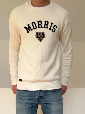 Vit tröja från Morris med brodyr - Vit stickad tröja från Morris med svart broderad text och ett snyggt emblem på bröstet. Tröjan har rund halsringning, ribbade muddar och är perfekt för en stilren look. Materialet känns mjukt och bekvämt mot huden.