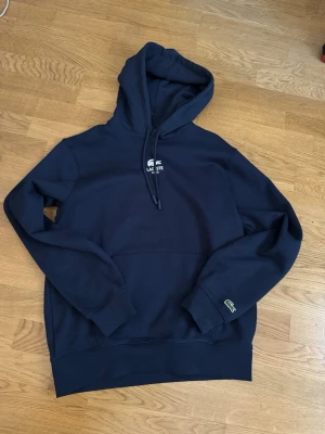 Mörkblå hoodie från Lacoste - Snygg mörkblå hoodie från Lacoste i unisexmodell med klassisk passform. Har huva med dragsko, broderad Lacoste-logga på bröstet och ärmslutet samt känguruficka framtill. Perfekt för en chill och stilren look.