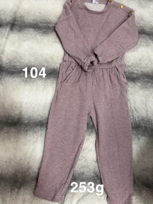 Lila/rosa jumpsuit med knappar - Mysig lila/rosa jumpsuit med långa ärmar och ben, resår i midjan och praktiska knappar på axlarna. Perfekt för en avslappnad och bekväm stil.🌸 Fläckar. Färgen är något mellan lila och rosa. Storlek 104