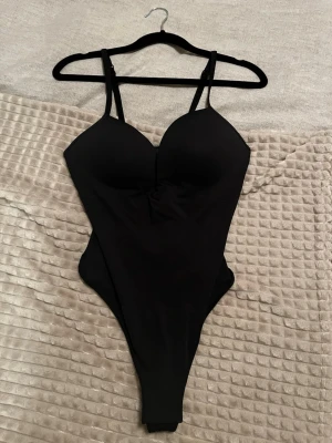 Svart body från H&M med vadderade kupor - Snygg svart body från H&M med vadderade kupor och justerbara axelband. Tillverkad i stretchigt material som sitter tight och formar kroppen. Perfekt för att skapa en smickrande siluett under kläderna.