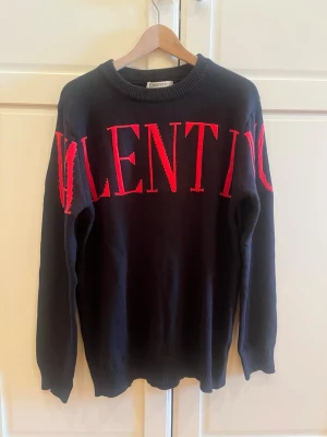 Svart stickad tröja från Valentino - Snygg svart stickad tröja från Valentino med stora röda bokstäver över bröstet och ärmarna. Tröjan har rund halsringning och långa ärmar. Perfekt för dig som gillar statement-plagg och exklusiva märken. Har ej kvitto men påse och etikett finns med 