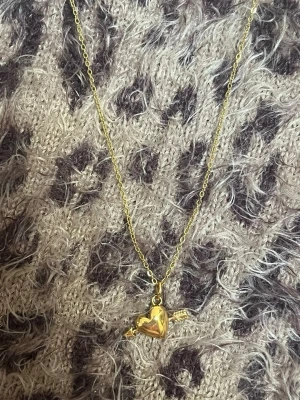 Guldigt halsband med hjärthänge litet  - Ett snyggt halsband i guldfärg med ett hjärthänge som har en pil genom sig. Kedjan är tunn och elegant, och hjärtat har en blank finish som ger ett coolt intryck. Perfekt för dig som gillar unika detaljer och vill ha något som sticker ut. Rostfri 