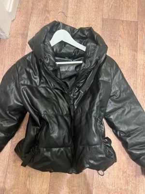 Svart pufferjacka med huva - Snygg svart pufferjacka i glansigt material med dragkedja framtill och två fickor med dragkedja. Jackan har en stor huva och justerbar dragsko i midjan för extra passform. Perfekt för kalla dagar när du vill hålla stilen.
