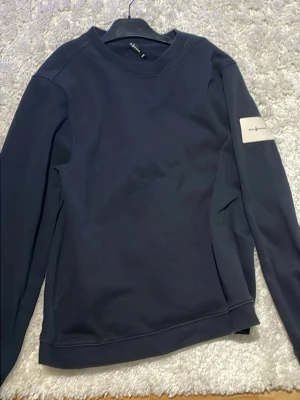 Mörkblå sweatshirt från Sail Racing - Mörkblå sweatshirt från sail racing med rund halsringning och lång ärm. Tröjan har en vit patch med logga på vänster ärm och är tillverkad i mjuk bomullsblandning. Enkel och stilren design som passar till det mesta.