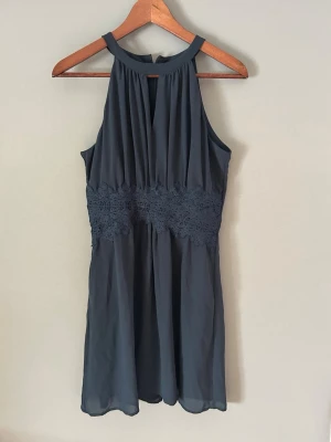 Mörkblå ärmlös klänning med spets - Snygg mörkblå kortklänning med spetsdetaljer i midjan och ärmlös design. Klänningen har hög halsringning och lätt, flödande tyg som ger en elegant siluett. Perfekt för dig som vill ha en stilren look med fina detaljer.