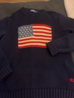 Mörkblå stickad tröja Ralph Lauren - Säljer en mörkblå stickad tröja från Ralph Lauren med en stor amerikansk flagga på bröstet. Tröjan har ribbstickade muddar vid ärmslut och nederkant samt rund halsringning. Perfekt för dig som gillar klassisk stil