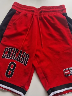 Röda Chicago basketshorts med tryck - Snygga röda basketshorts med svart och vit detalj, tryck med 'CHICAGO' och siffran 8 framtill. Elastisk midja med snörning och bakficka. Perfekta för sport eller streetwear.