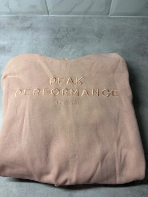 Beige/rosa hoodie från Peak Performance - Snygg beige hoodie från Peak Performance med broderad logga på bröstet. Mjuk insida och klassisk passform, perfekt för en avslappnad stil. Tillverkad i bomullsblandning för extra komfort.