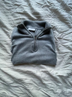 Quarter zip från GRUNT - Viktigt tröjan är i junior storlek.        Mysig grå stickad tröja med hög krage och halv dragkedja framtill. Tröjan har långa ärmar och ribbade muddar vid ärmslut och nederkant. Perfekt för lager-på-lager och enkel att matcha med det mesta.