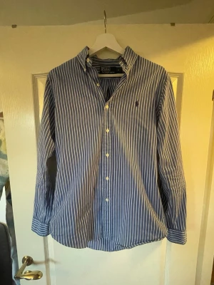 Blåvit randig skjorta från Polo Ralph Lauren - Klassisk blå skjorta med vita ränder från Polo Ralph Lauren. Skjortan har lång ärm, knappar framtill och en broderad logga på bröstet. Perfekt för en stilren och avslappnad look.