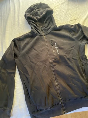Svart hoodie från Sail Racing - Svart hoodie med dragkedja från Sail Racing. Har huva med dragsko och en bröstficka med dragkedja och logga. 