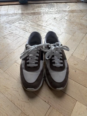 Arigato sneakers i vitt och brunt - Snygga sneakers från Axel Arigato i vitt tyg med bruna mockadetaljer och svart parti på sidan. Klassisk låg modell med vita snören och tjock sula för extra komfort. Perfekta för dig som vill ha en stilren och trendig look.