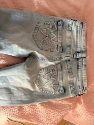 Ljusblå jeans från Gina tricot 14+ - Säljer ett par ljusblå bootcut jeans med snygga broderade bakfickor i vågmönster. Jeansen har låg midja och klassisk femficksdesign. Perfekta för dig som gillar en avslappnad och trendig stil.