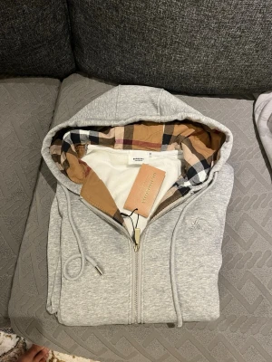 Grå zip-hoodie från Burberry - Helt ny snygg grå hoodie med dragkedja från Burberry. Hoodien har klassiskt rutigt foder i huvan, dragsko och två fickor framtill. Perfekt för en stilren och avslappnad look. Materialet är mjukt och bekvämt, och loggan är diskret broderad på bröstet. Storlek M passar även L