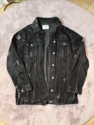 Svart distressed jeansjacka från Pull&Bear - Cool svart jeansjacka från Pull&Bear med slitna detaljer och klassisk knappstängning. Jackan har två bröstfickor med lock och knappar samt raka ärmar. 