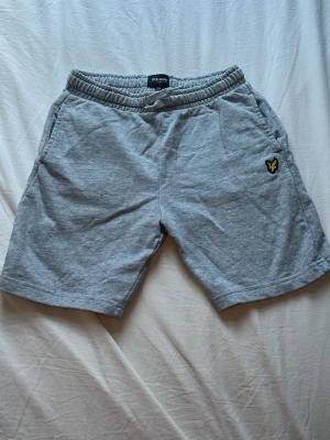 Grå shorts från Lyle & Scott - Grå mjuka shorts från Lyle & Scott med elastisk midja och snörning. Klassisk design med två sidofickor och den ikoniska gula loggan på vänster ben. Perfekta för en avslappnad stil under varma dagar. Endast använd tre gånger.