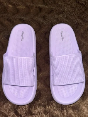 Ljuslila slides från HappyFlops - Säljer ett par ljuslila slides från HappyFlops. De har en bred, ribbad rem över foten och är gjorda i ett mjukt och flexibelt syntetmaterial. Perfekta att glida i hemma eller på stranden. Snygg minimalistisk design med öppen tå och räfflad sula för extra grepp.