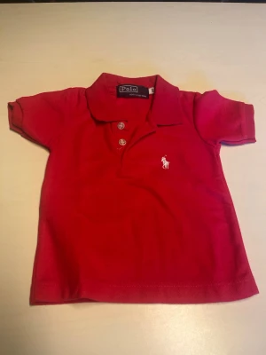 Röd pikétröja från Polo Ralph Lauren - Klassisk rosa pikétröja från Polo Ralph Lauren med vit broderad logga på bröstet. Tröjan har korta ärmar, krage och två knappar framtill. Perfekt för en stilren och sportig look.