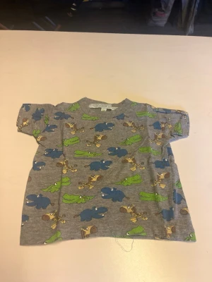 Grå t-shirt med djurmönster - Grå t-shirt med korta ärmar och färgglada tryck av flodhästar, giraffer och krokodiler. Perfekt för dig som gillar lekfulla och roliga motiv. Tillverkad i mjuk bomull som känns skön mot huden.
