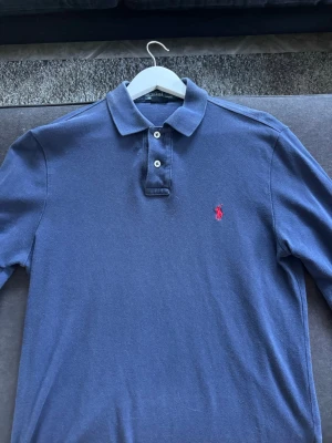 Marinblå långärmad piké från Ralph Lauren - Snygg marinblå långärmad pikétröja från Ralph Lauren med klassisk krage och två knappar framtill. Tröjan har det ikoniska röda broderade logotypen på bröstet och är tillverkad i mjuk bomull. Slimfit!
