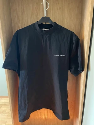 Svart t-shirt från Samsøe Samsøe - Svart t-shirt från Samsøe Samsøe med liten vit logga på bröstet. Klassisk rund halsringning och korta ärmar. Tillverkad i mjuk bomull. Aldrig använd.