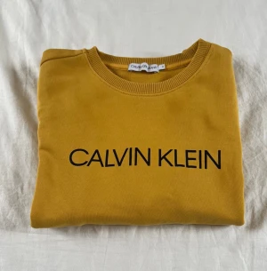 Gul sweatshirt från Calvin Klein - Snygg gul sweatshirt från Calvin Klein Jeans med svart logga framtill. Tröjan har rund halsringning och ribbade muddar. Perfekt för dig som vill sticka ut med en färgglad och trendig look.