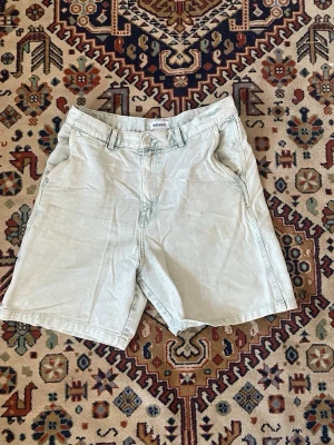 Ljusblå denimshorts från Weekday - Säljer ett par ljusblå denimshorts från Weekday med klassisk femficksdesign och bälteshällor. Shortsen har normal passform och stängs med dragkedja och knapp. Perfekta för varma dagar och enkel att matcha med olika stilar.