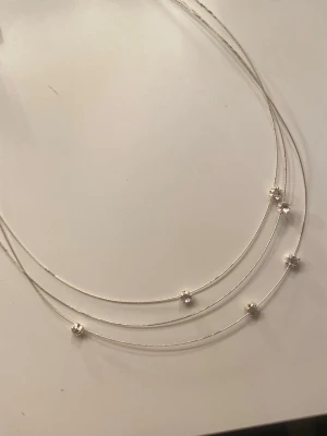 Tunt silverfärgat halsband med pärlor - Snyggt halsband i tre tunna silverfärgade kedjor med små vita pärlor placerade längs kedjorna. Halsbandet har ett justerbart lås med förlängningskedja, perfekt för att anpassa längden. Stilren och modern design som passar till många outfits. Helt oanvänd 