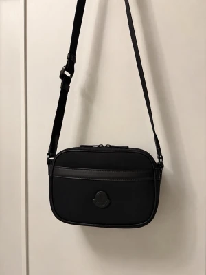 Svart axelväska från Moncler - Snygg svart axelväska från Moncler med justerbar rem och minimalistisk design. Väskan har en oval form, diskret Moncler-logga framtill och detaljer i svart läder. Perfekt för dig som gillar stilrena accessoarer och vill ha plats för det viktigaste.