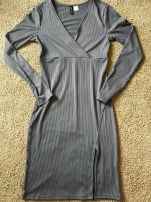Grå ribbad klänning från H&M Divided - Snygg grå ribbad klänning från H&M Divided med v-ringning och långärmad design. Klänningen har en slits framtill och är figurnära med kort längd. Perfekt för dig som gillar stilrena och enkla plagg med en modern touch.