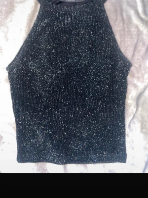 Glittrig svart polo  topp - Supersnygg svart glittrig topp med hög hals och ärmlös design. Perfekt för dig som vill sticka ut med lite extra glitter. Toppens material är stretchigt och sitter tajt mot kroppen.