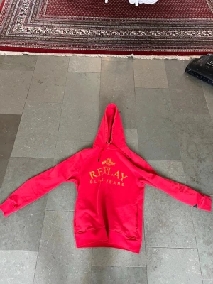 Röd Replay hoodie med tryck - Snygg röd hoodie från Replay med gul text 'REPLAY BLUE JEANS' och emblem på bröstet. Tillverkad i mjuk ekologisk bomull och har en klassisk huva samt känguruficka framtill. Perfekt för en avslappnad och trendig stil.