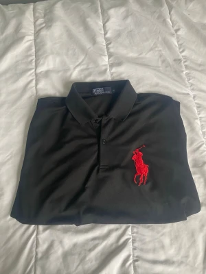 Svart pikétröja från Polo Ralph Lauren - Svart pikétröja från Polo Ralph Lauren med stor röd broderad logga på bröstet. Klassisk krage och knappar framtill. Perfekt för en stilren och sportig look.
