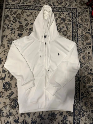 Vit hoodie från Polo Ralph Lauren - Snygg vit hoodie med dragkedja från Polo Ralph Lauren. Klassisk design med svart broderad logga på bröstet, huva med dragsko och ribbade muddar. Perfekt för en clean och stilren look. Materialet känns mjukt och bekvämt.